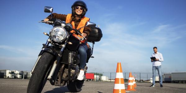 Student at motorcycle driving school with helmet taking motorcycle lessons and practicing riding. In the background, there are traffic cones and an instructor with checklist rating and evaluating the ride.