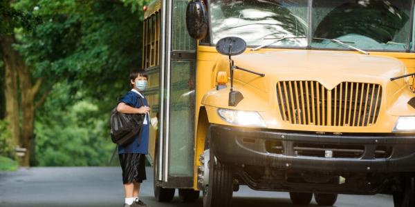 School bus picking up elementary student wearing surgical mask boarding at bus stop.